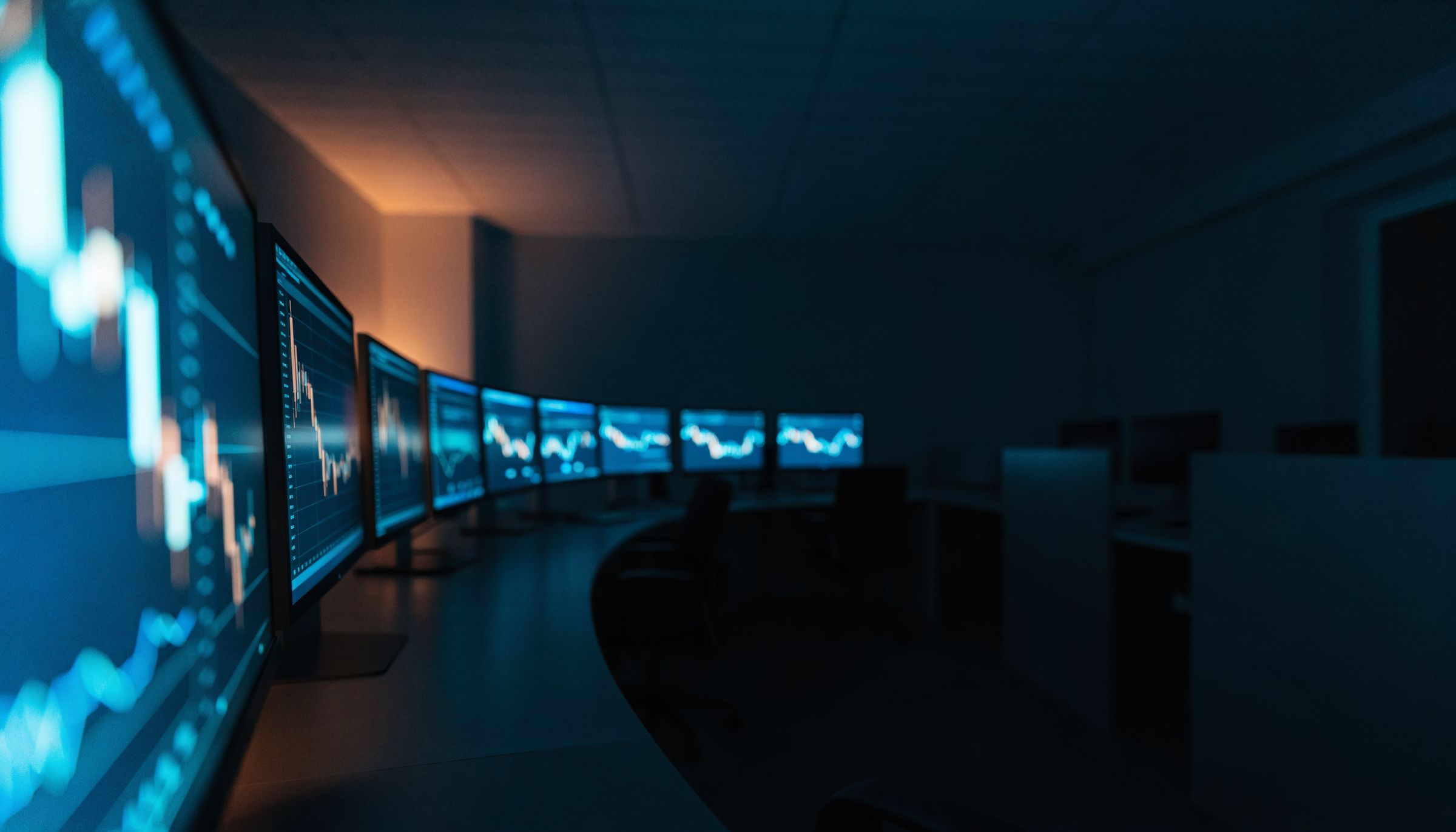 A darkened trading floor, its curved bank of monitors glowing with price charts.